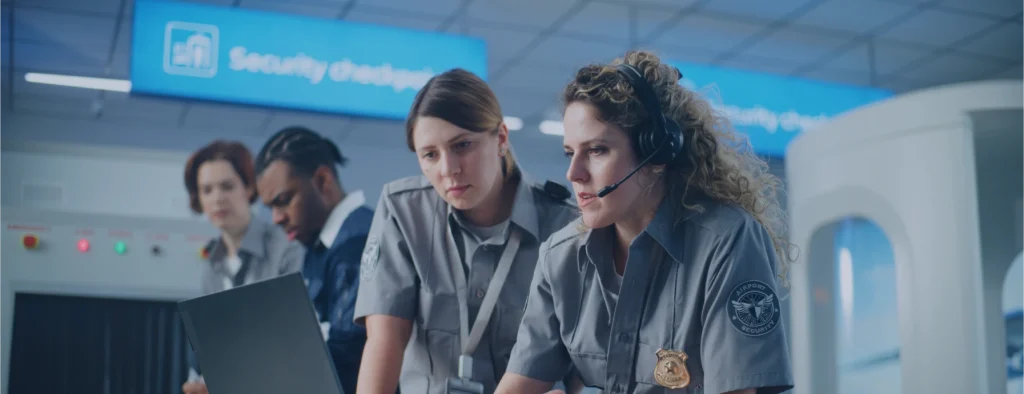Airport security staff gather at a checkpoint workstation, reviewing information on a laptop beneath signage for security check-in.