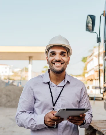 Construction worker wearing a hard hat holds a tablet at an outdoor work site with a truck and dirt piles in the background.