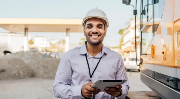 Person wearing a hard hat uses a tablet at a construction site beside large equipment.