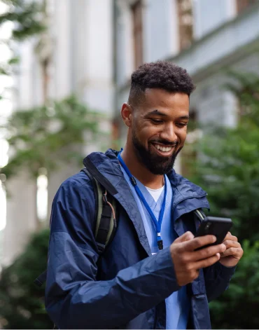 : Person wearing a backpack looks at a smartphone outdoors on a sidewalk near buildings and greenery.