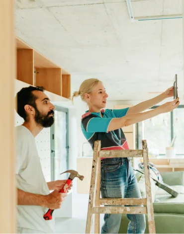 Two people work indoors on a home renovation, with one person standing on a ladder holding a measuring tool while another holds a hammer nearby.