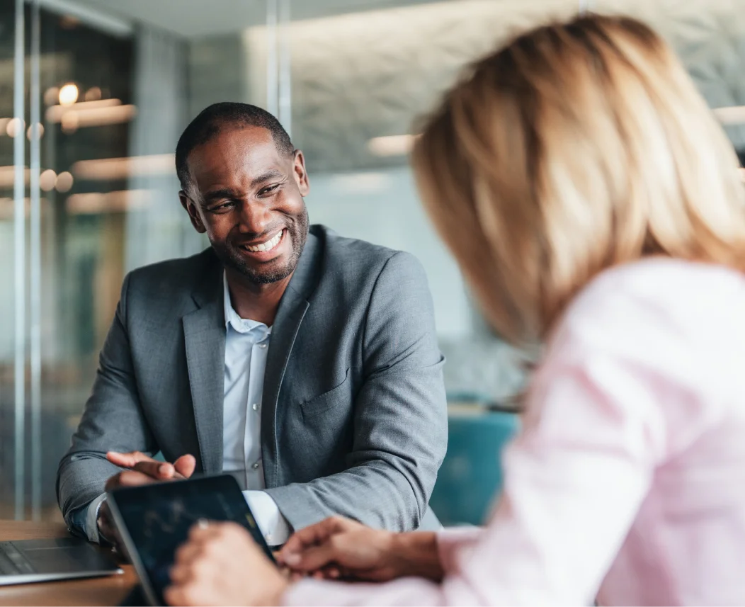 Two people meet at a table in a modern office, with one person holding a tablet.