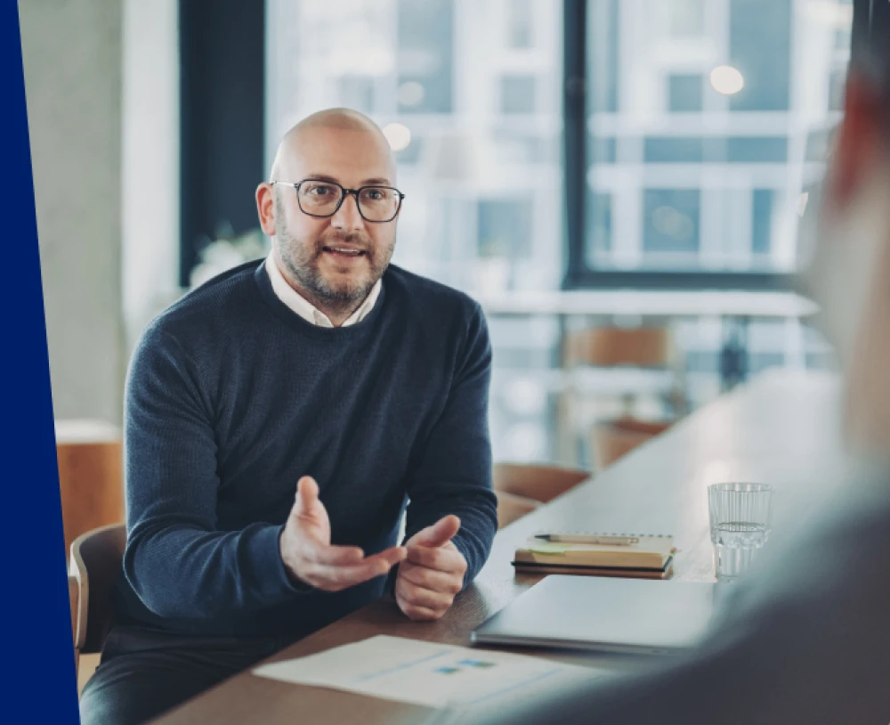 A banker sits at a table in a bright office setting during a conversation, with documents, a notebook, and a laptop nearby.