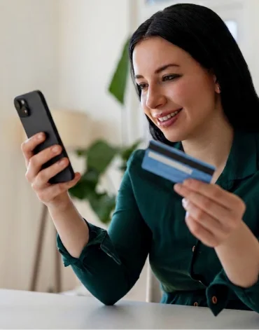 Person holding a smartphone and a credit card while looking at the phone, seated indoors with a softly blurred home background.