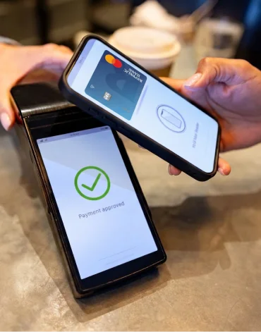 A person uses a smartphone to make a contactless payment at a card reader displaying “Payment approved.”