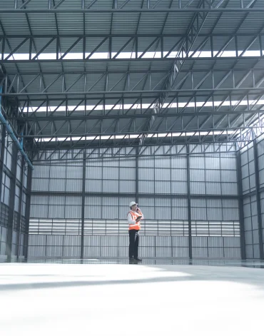 A worker wearing a safety vest stands inside a large, empty industrial warehouse.
