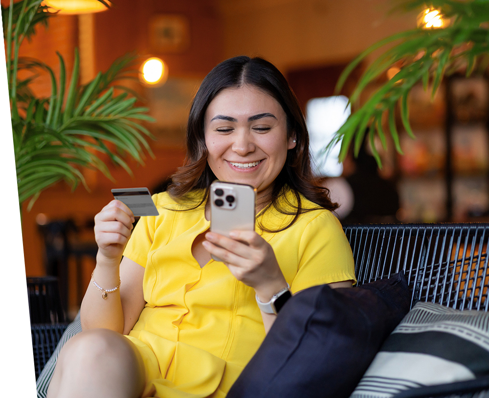 A person in a yellow outfit uses a smartphone while holding a bank card in a café.
