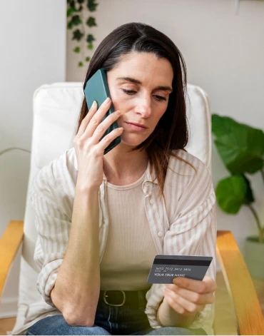 A person holds a bank card while speaking on a smartphone in a home setting.
